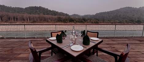 A dining table set for four with chairs, positioned on a wooden deck overlooking a river and hills.