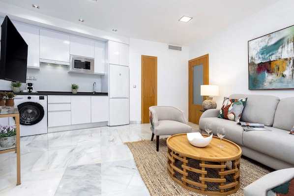A modern kitchen with white cabinets, a microwave, and a washing machine. There is a round wooden table with a white bowl and two glasses. A grey sofa and a chair are in the living area, with a painting on the wall. - Cozy 2BD Apartment in Cordoba Uceda I (Córdoba)
