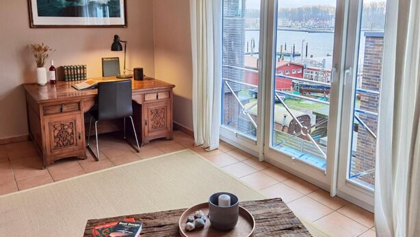 Een kamer met een bureau, stoel en uitzicht op een haven door grote ramen.