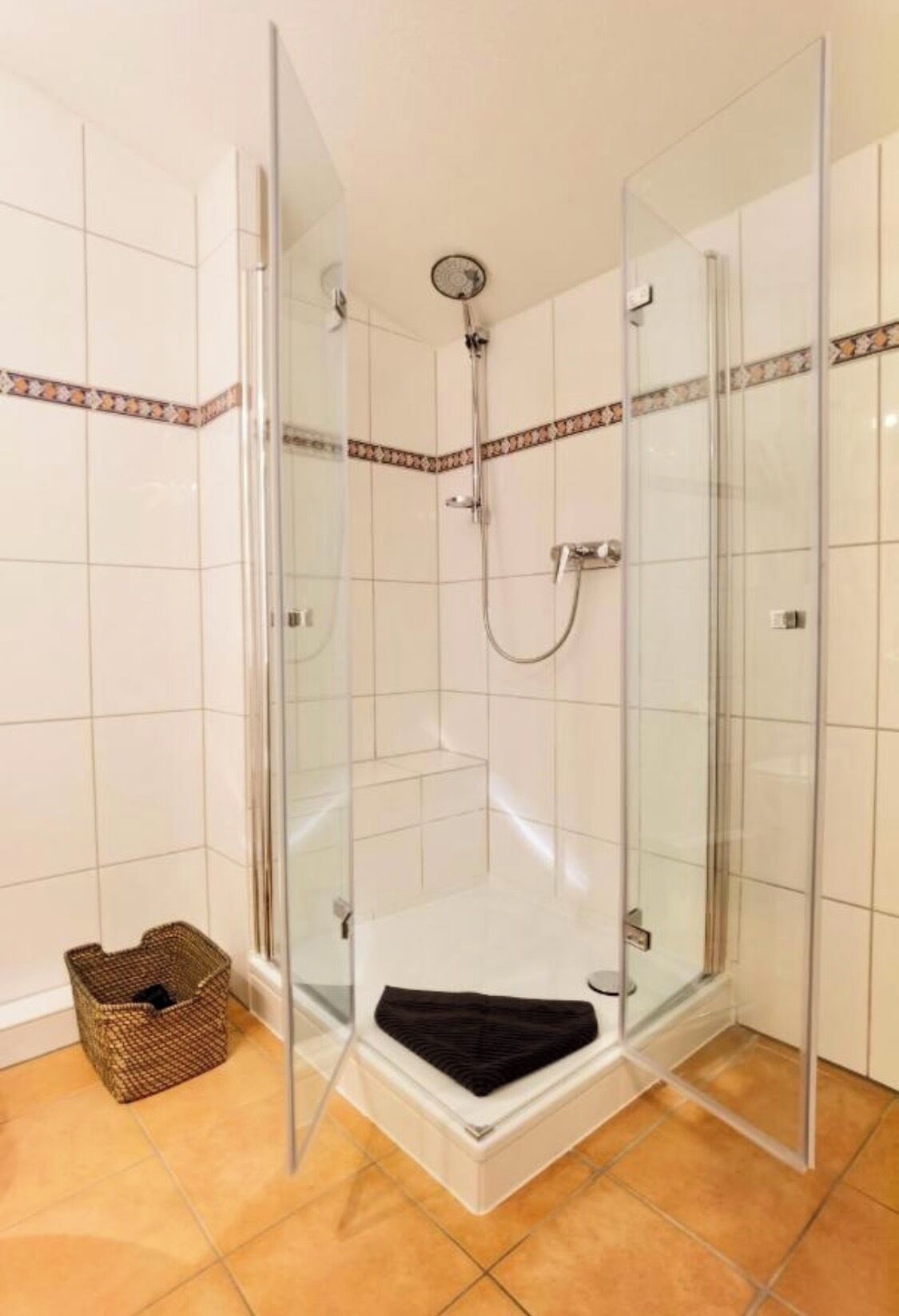 Cabina de dutxa amb vidre, dutxadora i mànega de dutxa, parets de rajola blanca i un estri de fons a la cantonada.