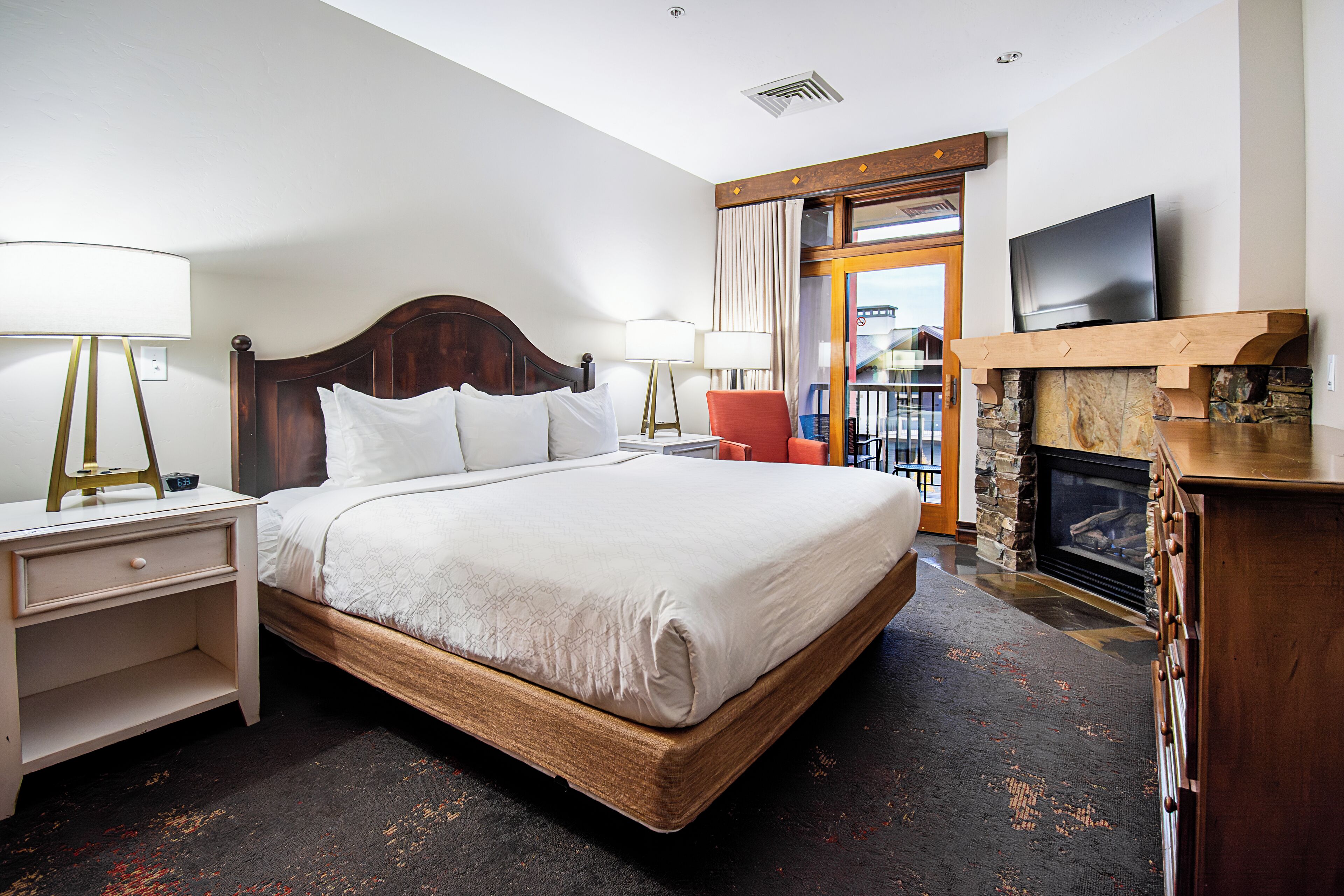 A hotel room with a large bed, two bedside tables with lamps, a television mounted above a fireplace, and a view of the outside through a glass door.