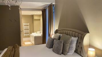 A hotel room with a bed, a chandelier, a bathroom visible through an open door, and a bedside lamp.