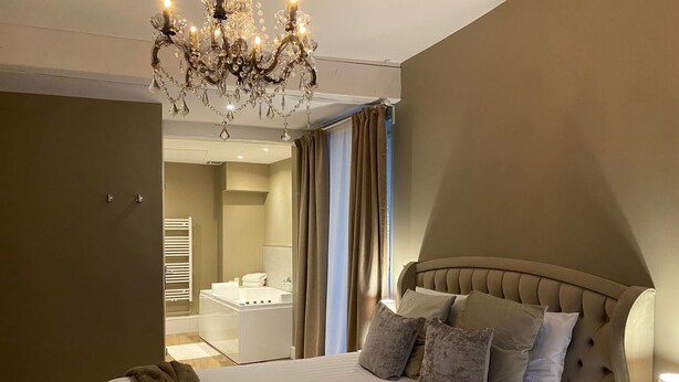 Une chambre avec un grand lit, un lustre, une salle de bain avec baignoire et une vue sur l’extérieur par une fenêtre.