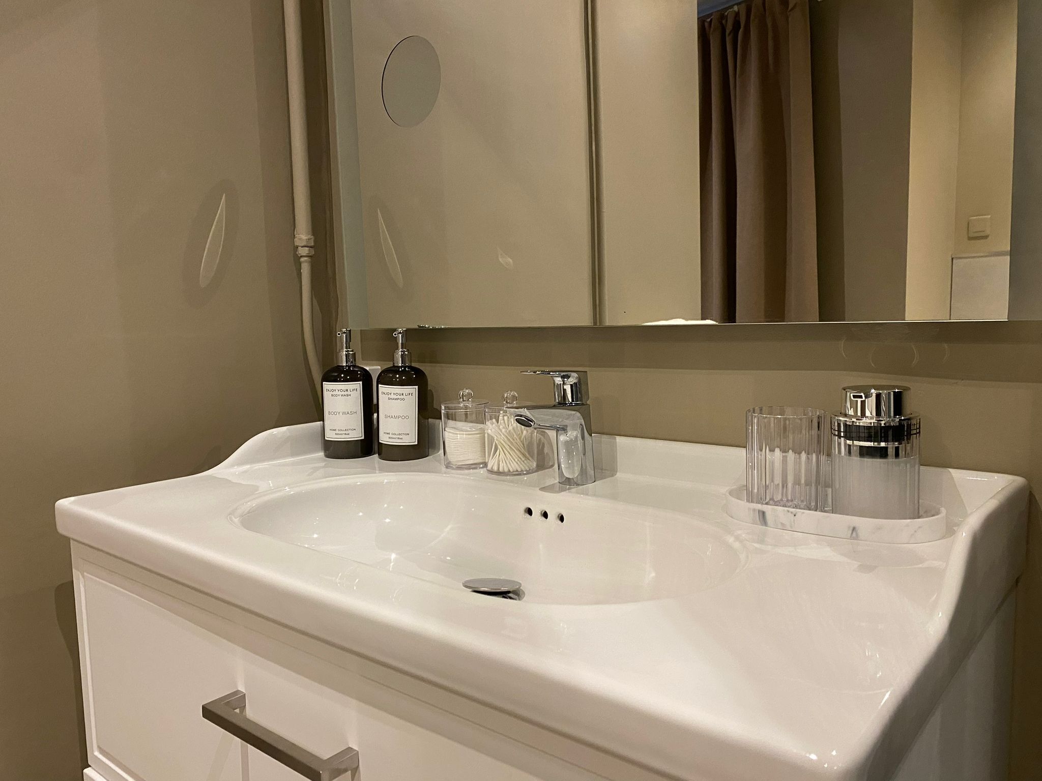 A bathroom with a white sink, two bottles of body wash and shampoo, a mirror, and a glass container with cotton swabs.