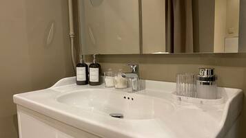 A bathroom with a white sink, two bottles of body wash and shampoo, a mirror, and a glass container with cotton swabs.