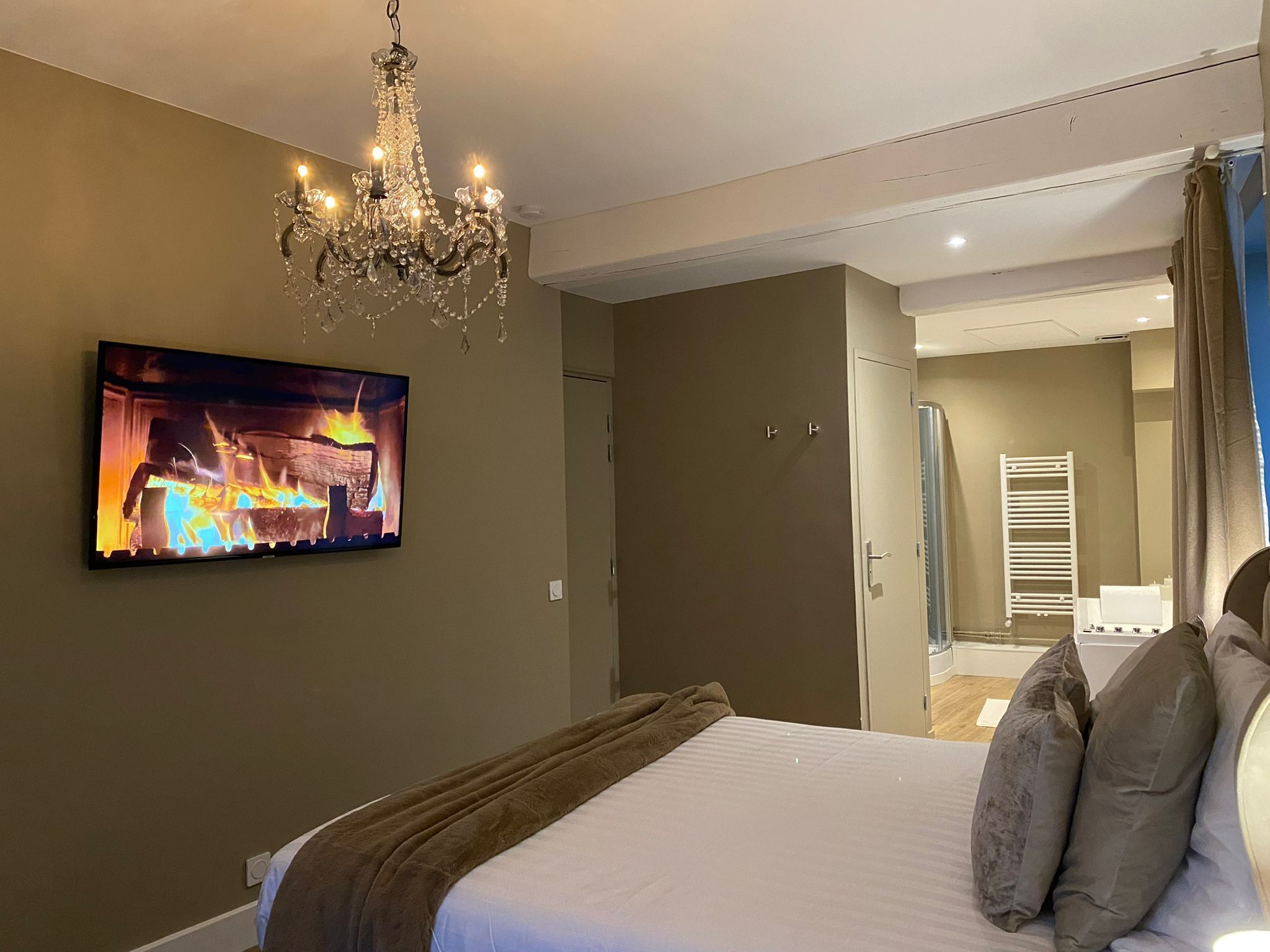 A bedroom with a large bed, a chandelier, a flat-screen TV, and a bathroom visible through an open door.