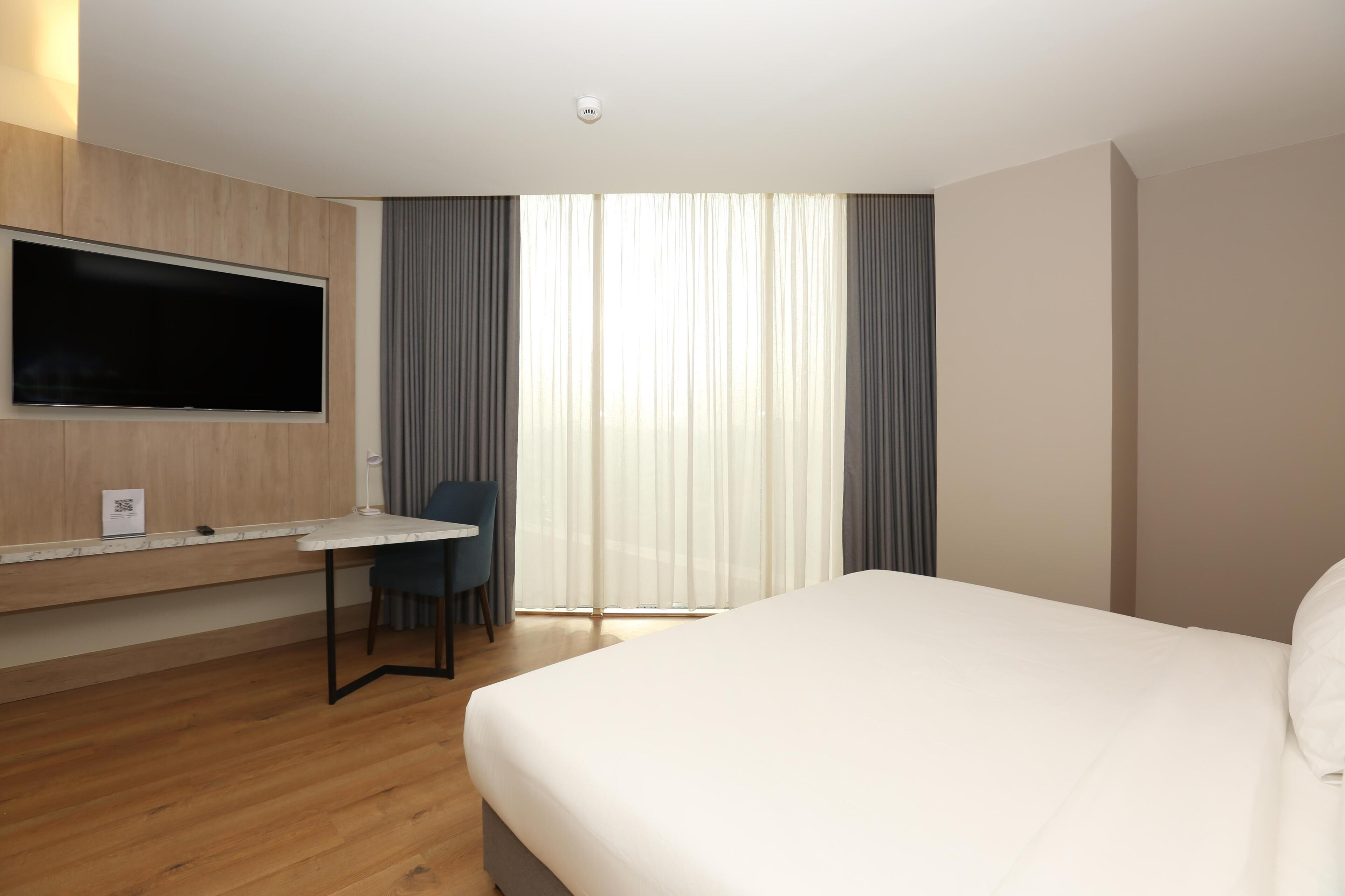 A hotel room with a bed, a desk, a chair, a flat-screen TV, and large windows with curtains.