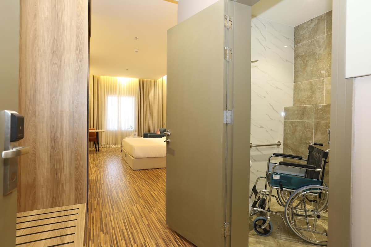 A hotel room with a wooden floor, a bed, a wheelchair, and a shower area.