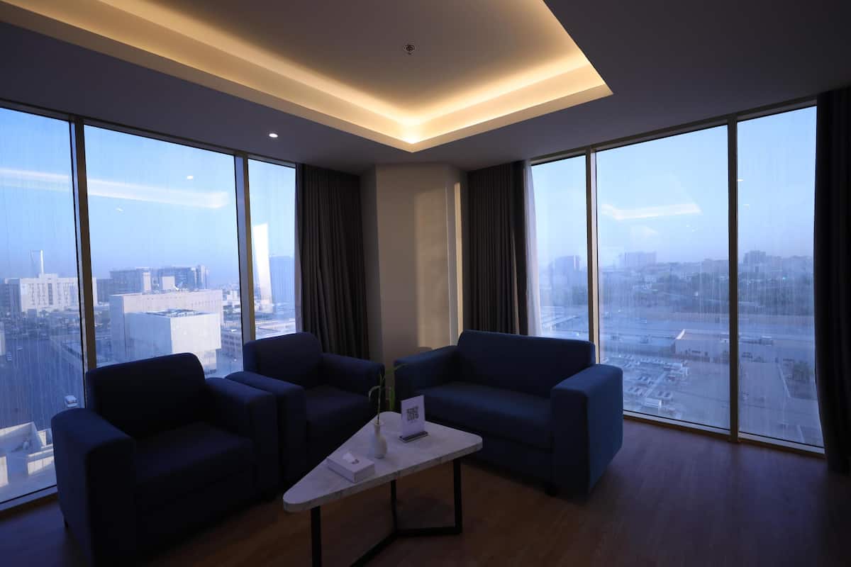 A modern hotel room with a large city view, two sofas, a coffee table, and a ceiling light fixture.
