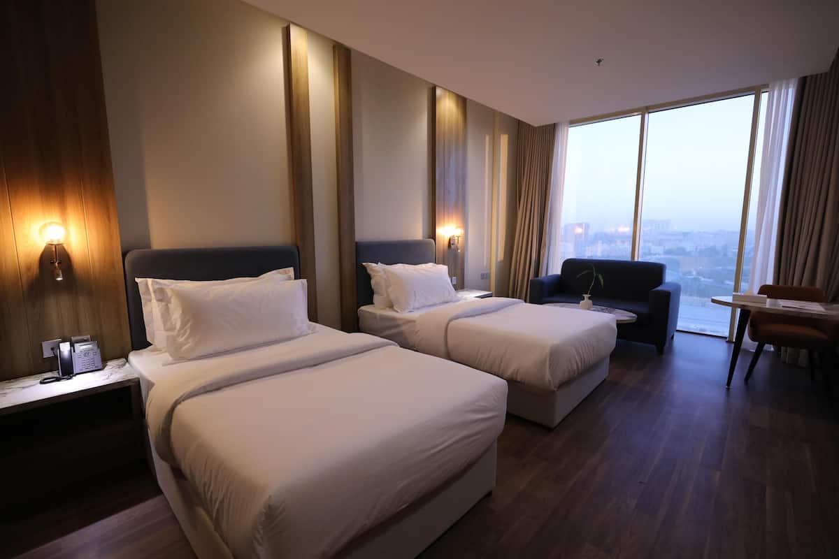 A hotel room with two beds, a sofa, a dining table, and a large window with a city view.