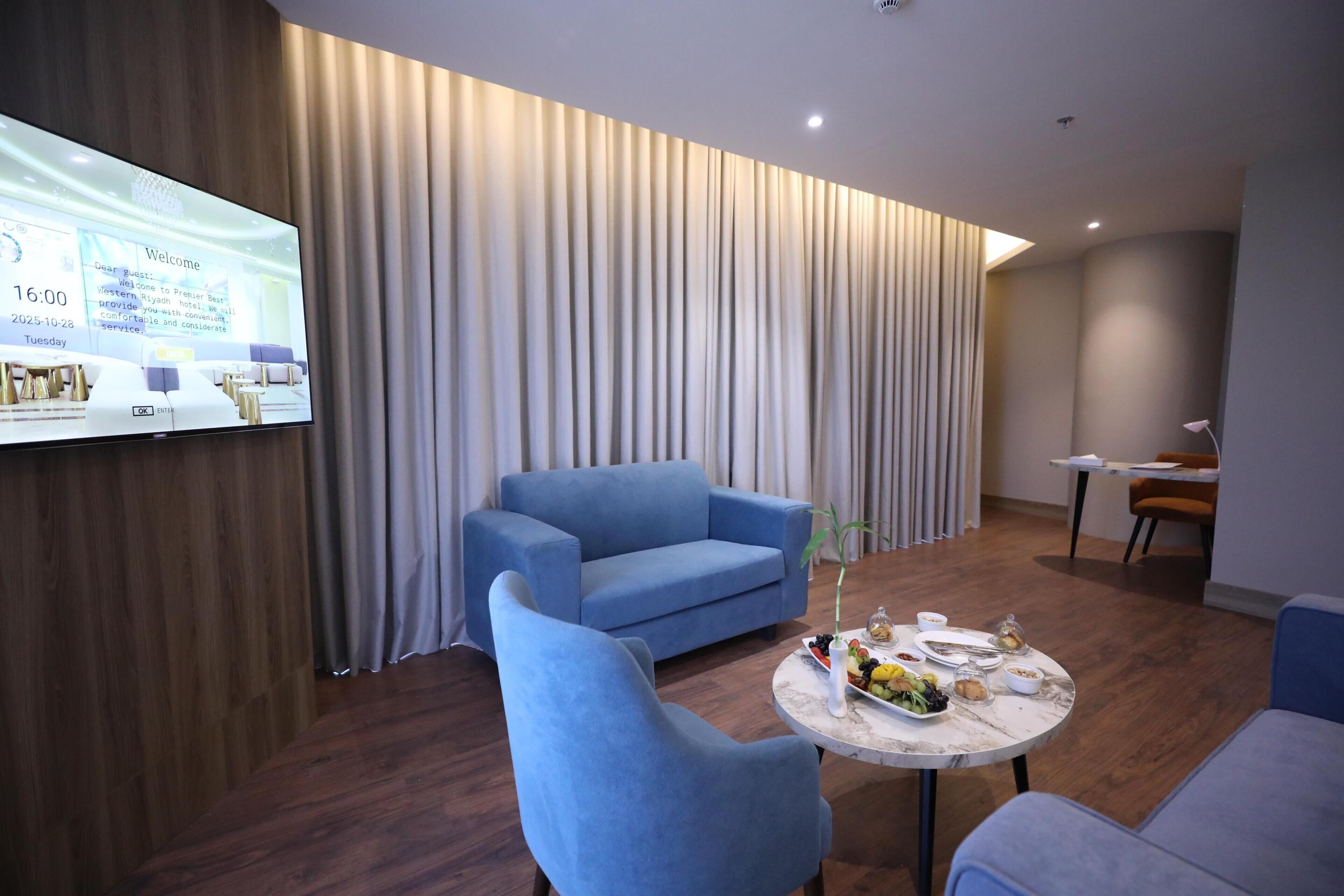 Habitación de hotel moderna con un televisor de pantalla plana grande, un sofá azul, una mesa redonda con un florero y suelo de madera.