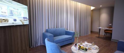 Habitación de hotel moderna con un televisor de pantalla plana grande, un sofá azul, una mesa redonda con un florero y suelo de madera.