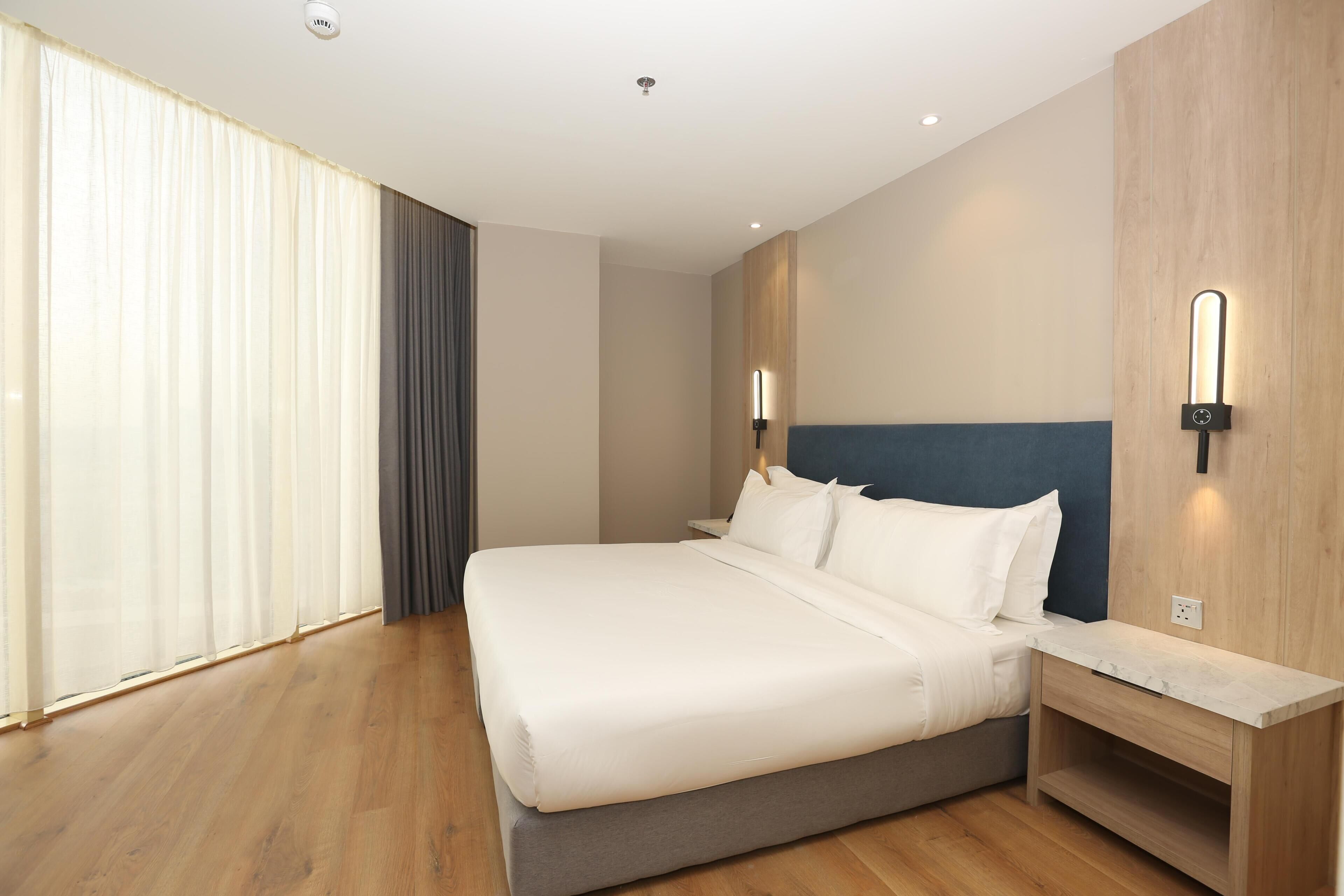 A hotel room with a large bed, wooden flooring, a nightstand, and a wall-mounted light fixture.