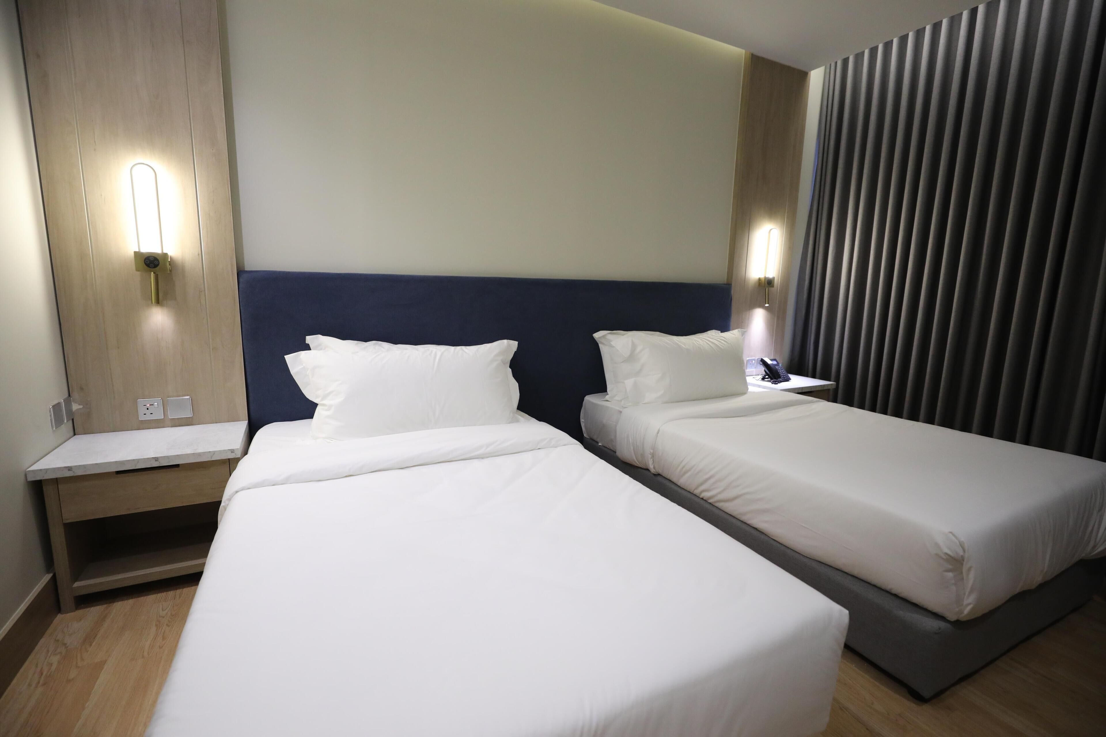 A hotel room with two single beds, a wooden nightstand, a wall-mounted light, and a curtain.