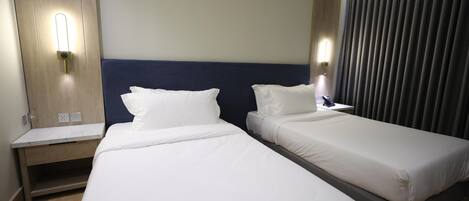 A hotel room with two single beds, a wooden nightstand, a wall-mounted light, and a curtain.