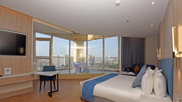A modern hotel room with a large bed, a desk, and a balcony with a city view.