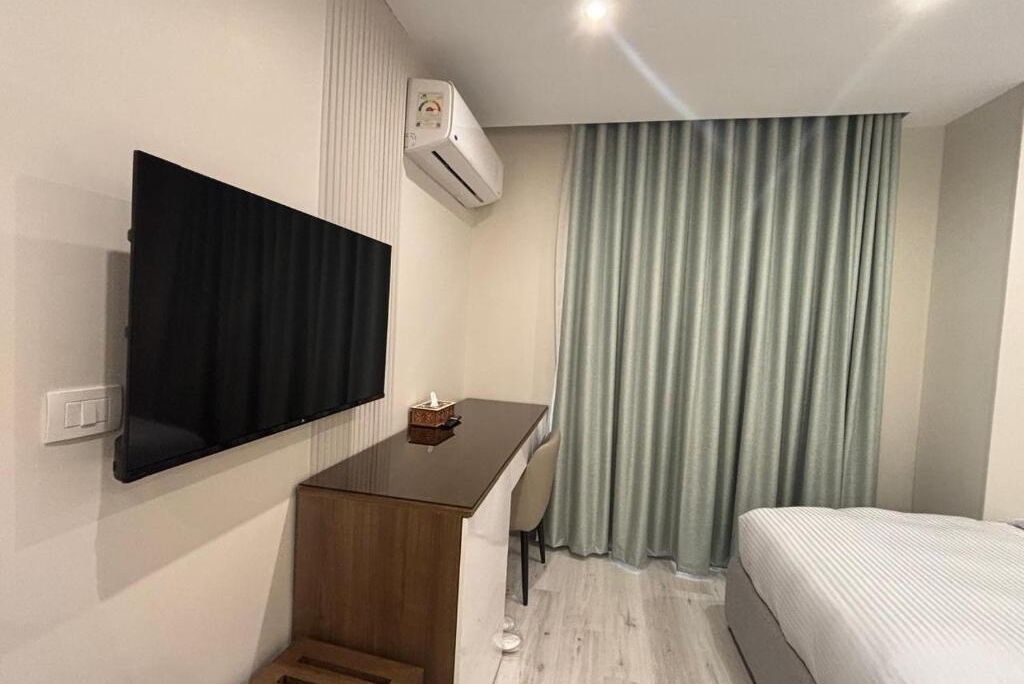 Une chambre d’hôtel comprenant un lit, un bureau avec une chaise, un téléviseur fixé au mur et une fenêtre avec des rideaux.