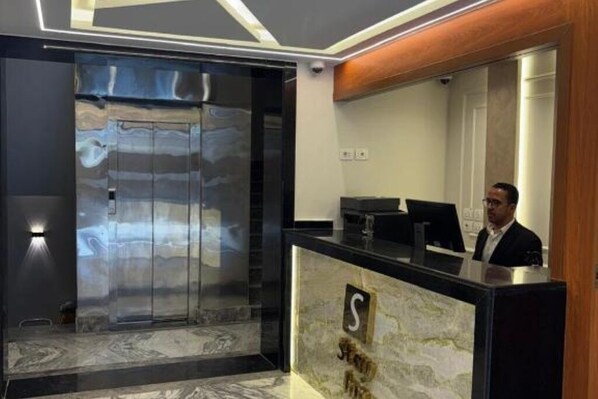 Un área de recepción con mostrador de mármol, un ascensor de vidrio y un panel de madera en la pared.