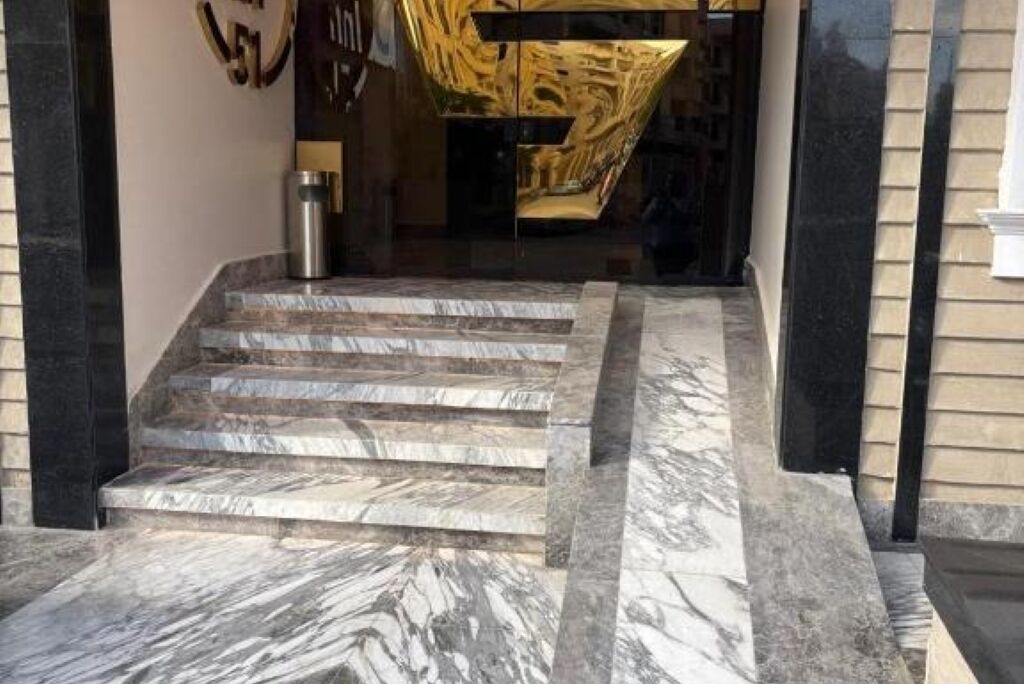 A set of marble steps leading to a building entrance with a glass door and a golden abstract sculpture above it.