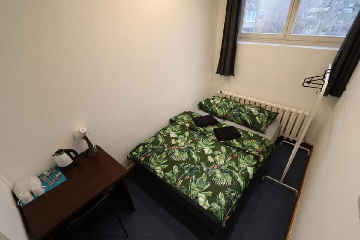 A small, compact room with a single bed featuring a tropical leaf pattern, a bedside table with a kettle and cups, and a radiator.