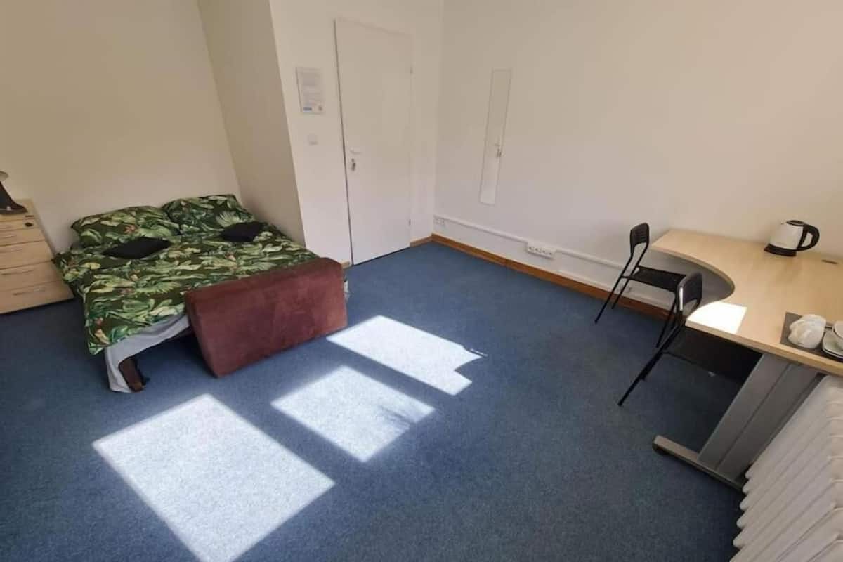 A small, sparsely furnished room with a bed, a desk, and a chair.