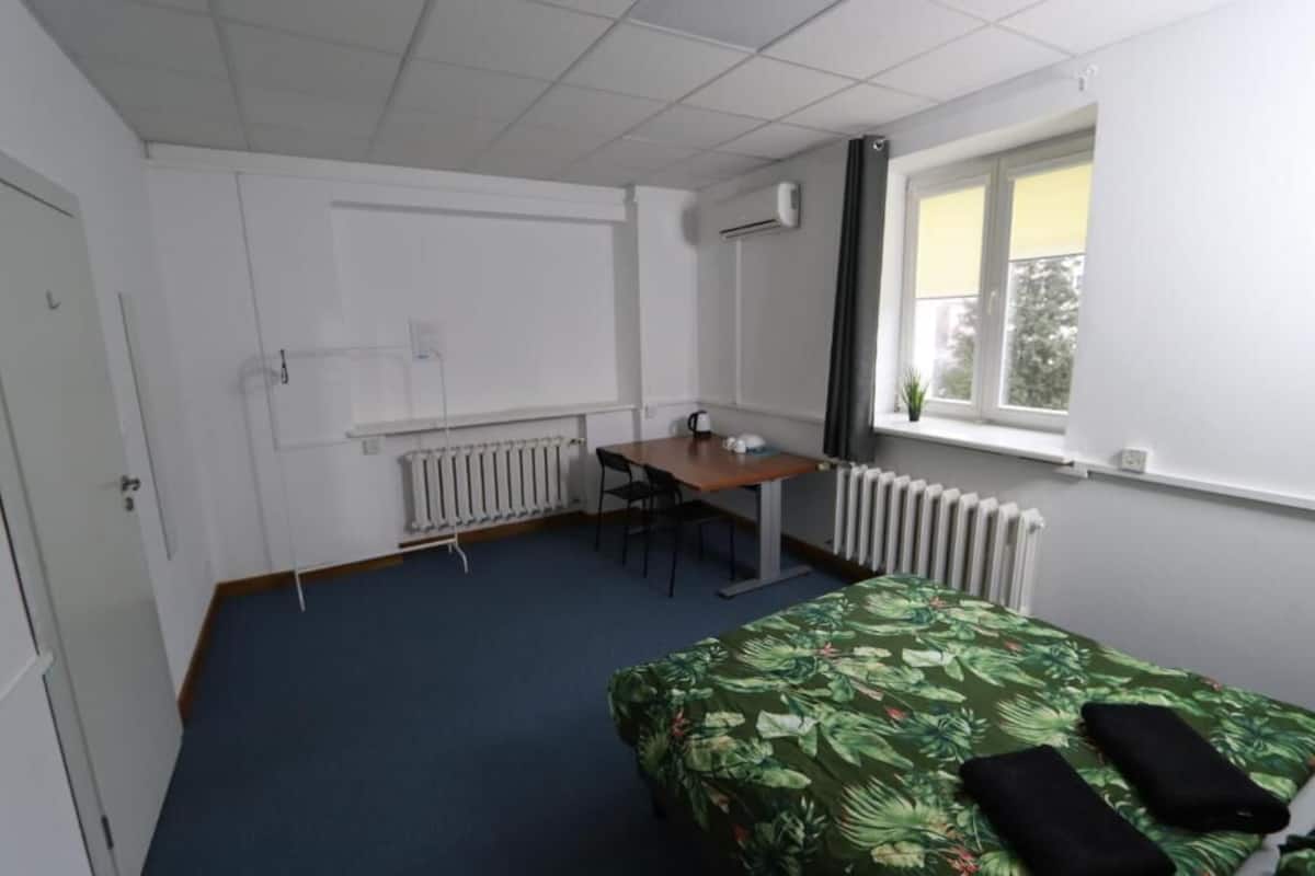 A small, clean room with a bed, a desk, and a chair. There is a window with a view of trees and a radiator under the window.