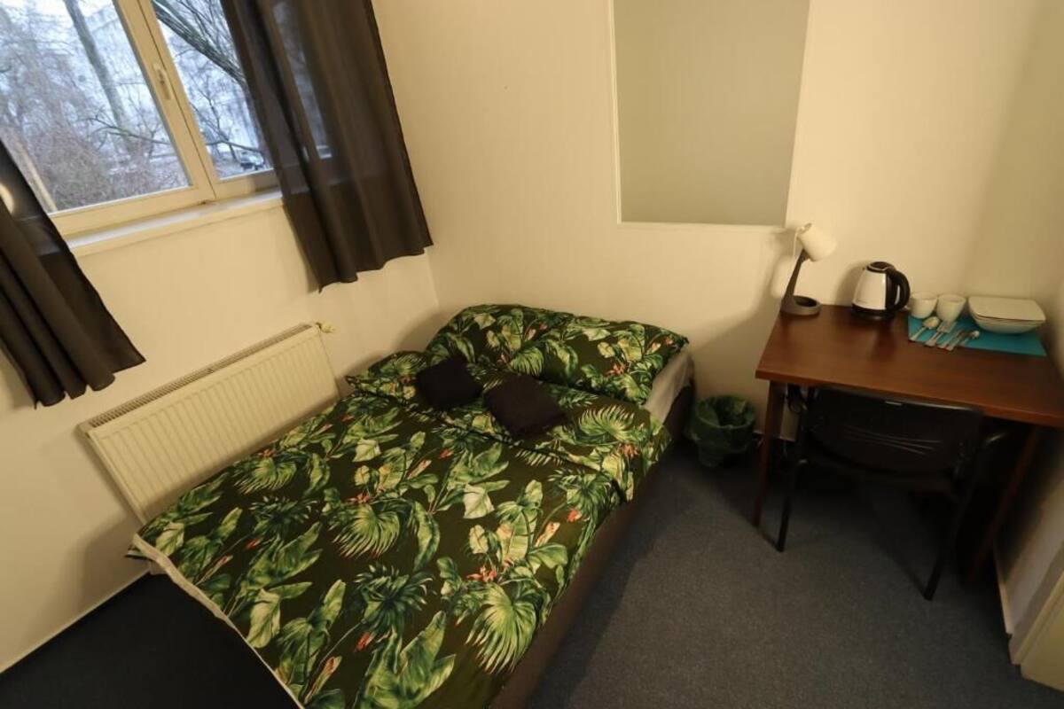 A small, single-bed room with a tropical leaf-pattern bedspread, a desk with a chair, a radiator, and a window with curtains.