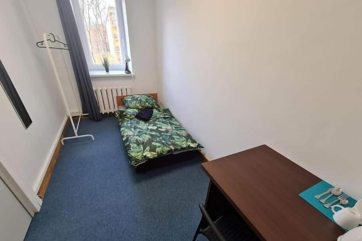 A small, tidy room with a single bed, a wooden desk, and a window with a view of buildings outside.