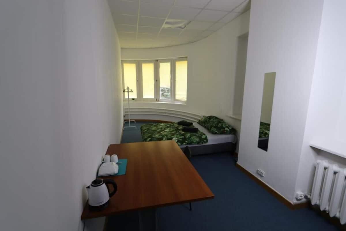 A small, compact room with a wooden table, a kettle, and a bed with green patterned bedding.