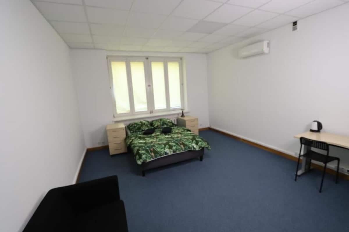A bedroom with a bed, a desk, a chair, a window, and an air conditioning unit.