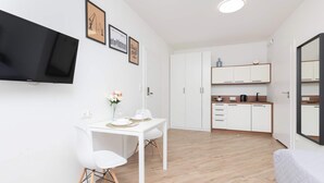 A modern, compact room with a white desk, chair, and bed. It features a flat-screen TV mounted on the wall, a small kitchen area with cabinets, and a mirror. - Modern Studio in Wrzeszcz | Balcony | Close to the Station (Gdańsk)