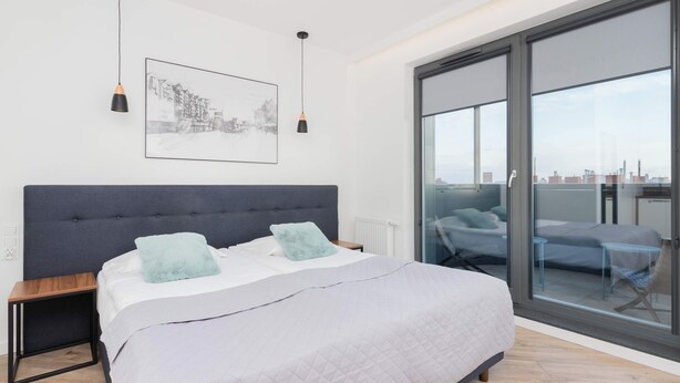 A modern bedroom with a large bed, a cityscape view through floor-to-ceiling windows, and a small table with a chair. - Modern Studio in Wrzeszcz | Balcony | Close to the Station (Gdańsk)