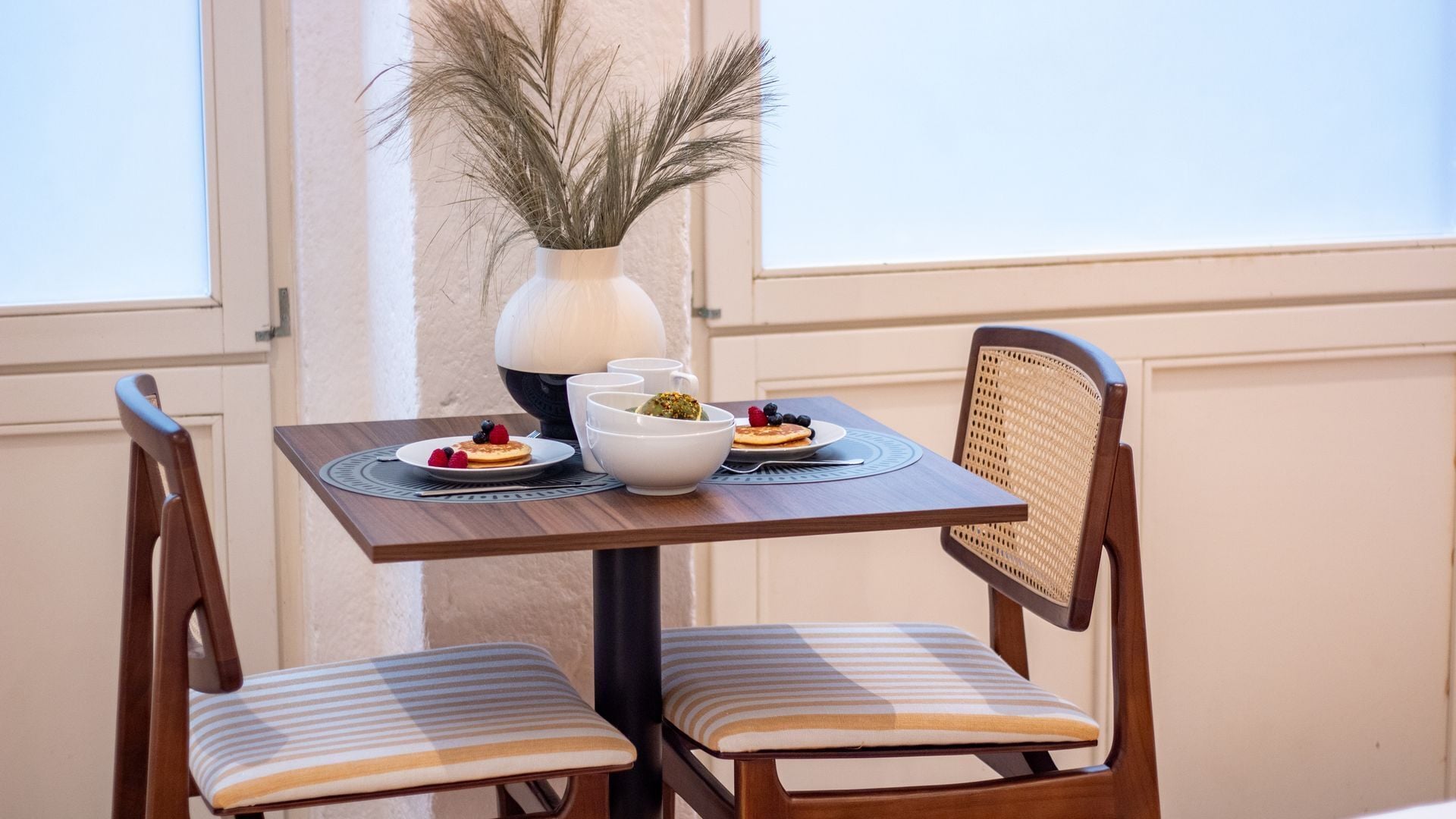 Una mesa de comedor con un jarrón de plantas secas, dos platos de comida y dos sillas a rayas.