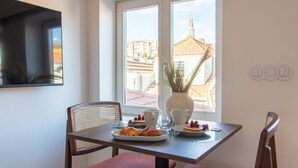 Une table à manger avec une assiette de pâtisseries et un vase contenant des plantes séchées, une chaise et une fenêtre donnant sur des immeubles.