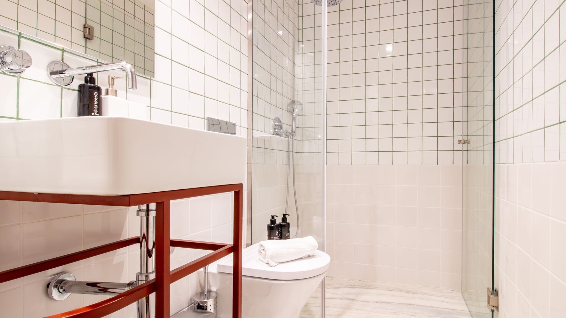 Un baño moderno con ducha de azulejos blancos, zona de ducha con cerramiento de vidrio, inodoro blanco y tocador con espejo de estructura de madera.