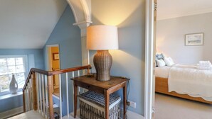 Una habitación con una mesa de madera, una lámpara, una cesta de mimbre y una cama con ropa blanca.