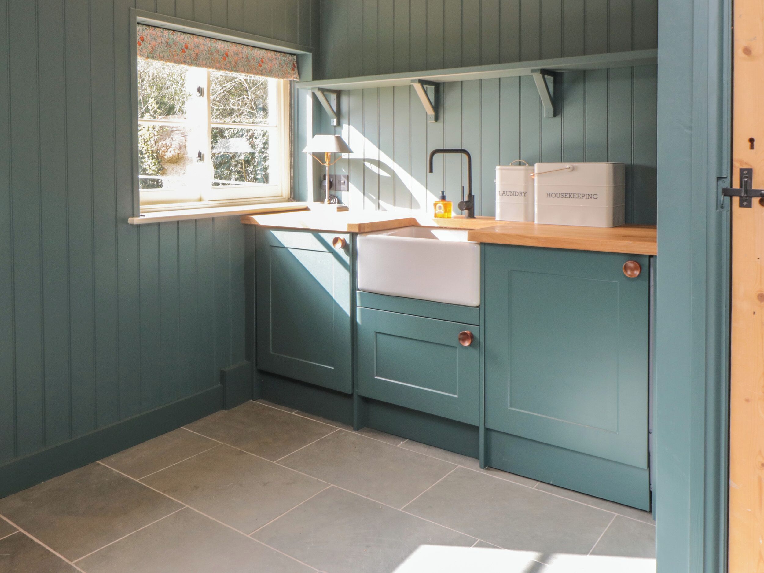 Een keuken met blauwgroene kastjes, een houten aanrechtblad en een raam met bloemen gordijn.