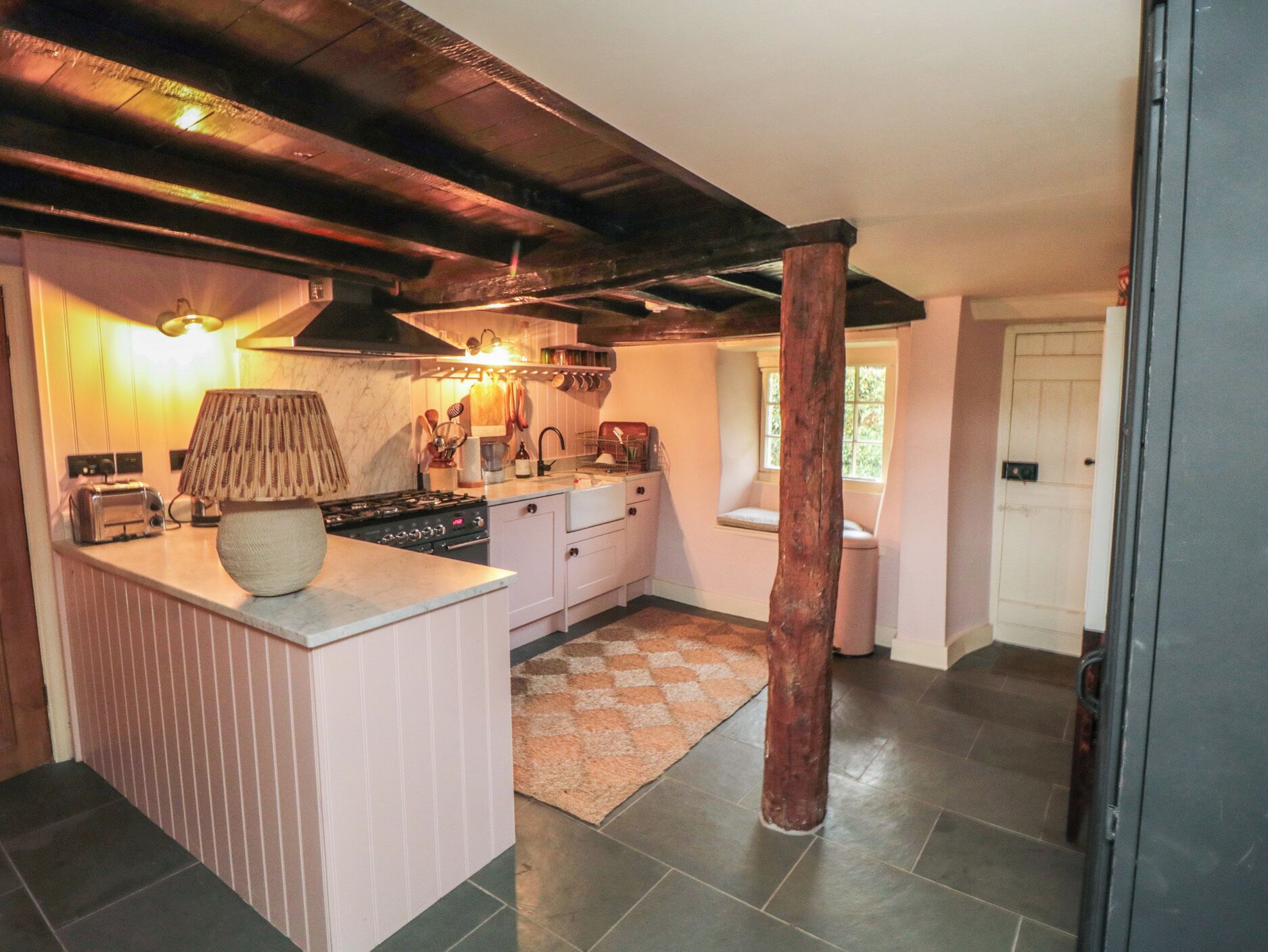 A kitchen with a central island, a stove, and a sink. There is a wooden beam ceiling and a window with a view of greenery.