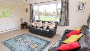 A living room with a dark brown leather sofa, a blue patterned rug, and a window with curtains. - Cherry Trees (Haverfordwest)