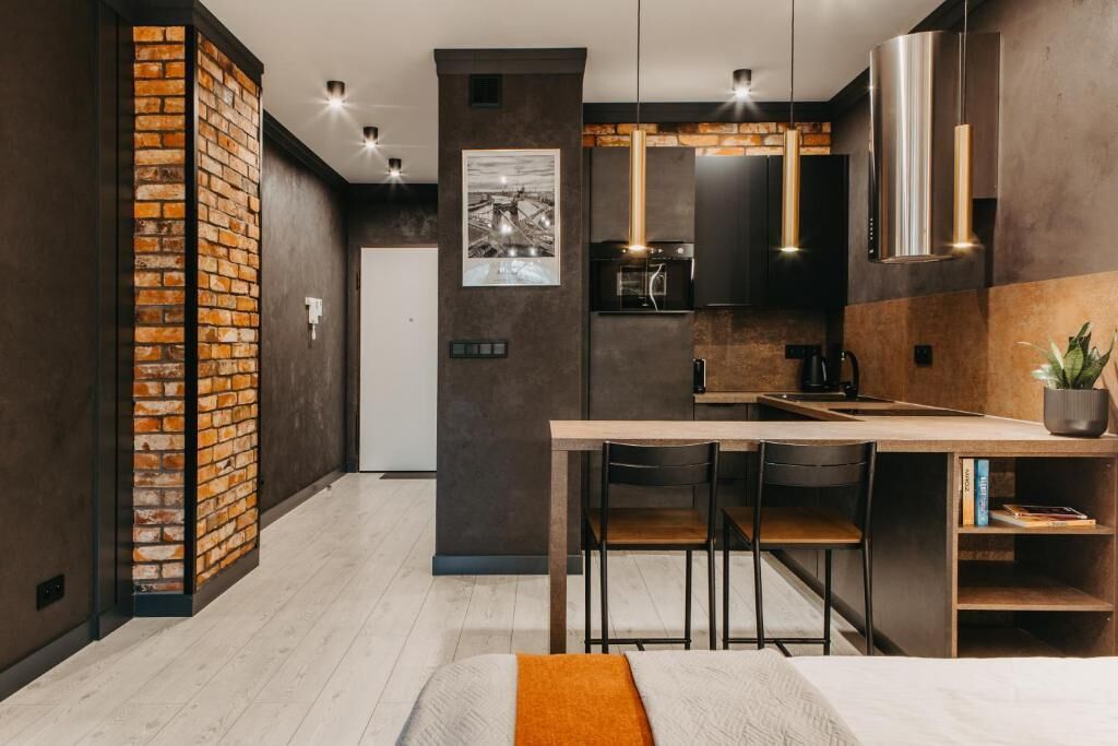 A modern kitchen with a brick accent wall, a dining area with a wooden table and chairs, and a bed with an orange throw.