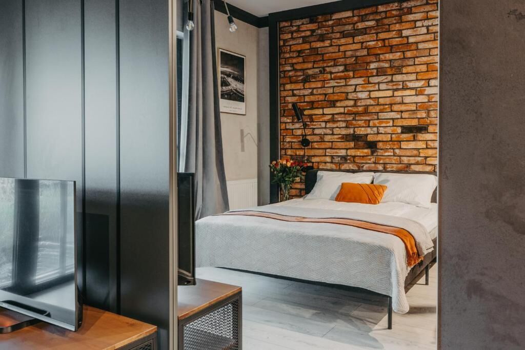 A modern bedroom with a brick accent wall, a bed with white bedding and orange pillows, a TV on a wooden stand, and a small side table.