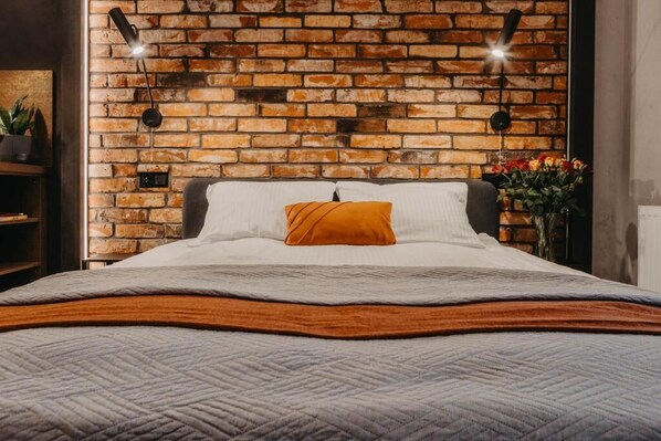 Una cama con cubrecolcha acolchado, una sola almohada naranja y una pared de ladrillo al fondo.