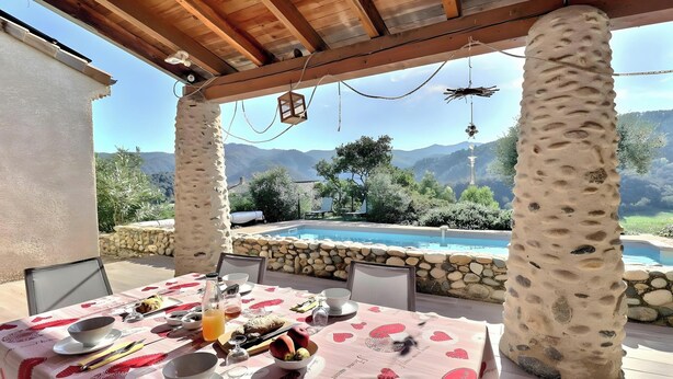 Un espace extérieur abrité, aménagé pour les repas, avec vue sur la piscine, des piliers en pierre et une table dressée.