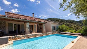 Une piscine bordée d’un muret en pierre et une maison avec un toit en tuiles.