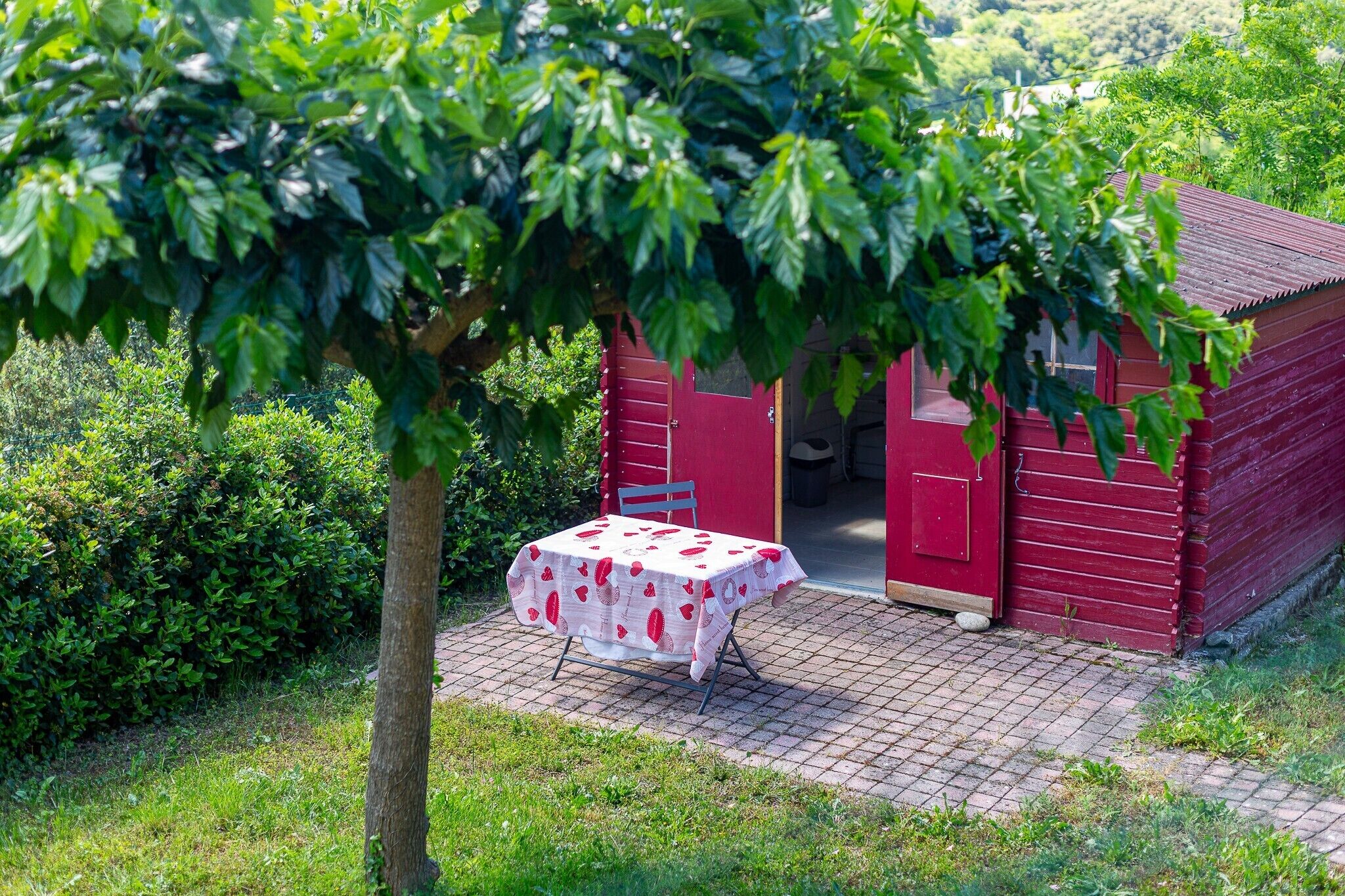 Un bâtiment en bois rouge, avec une table recouverte d’une nappe blanche ornée de motifs rouges, le tout entouré de verdure.