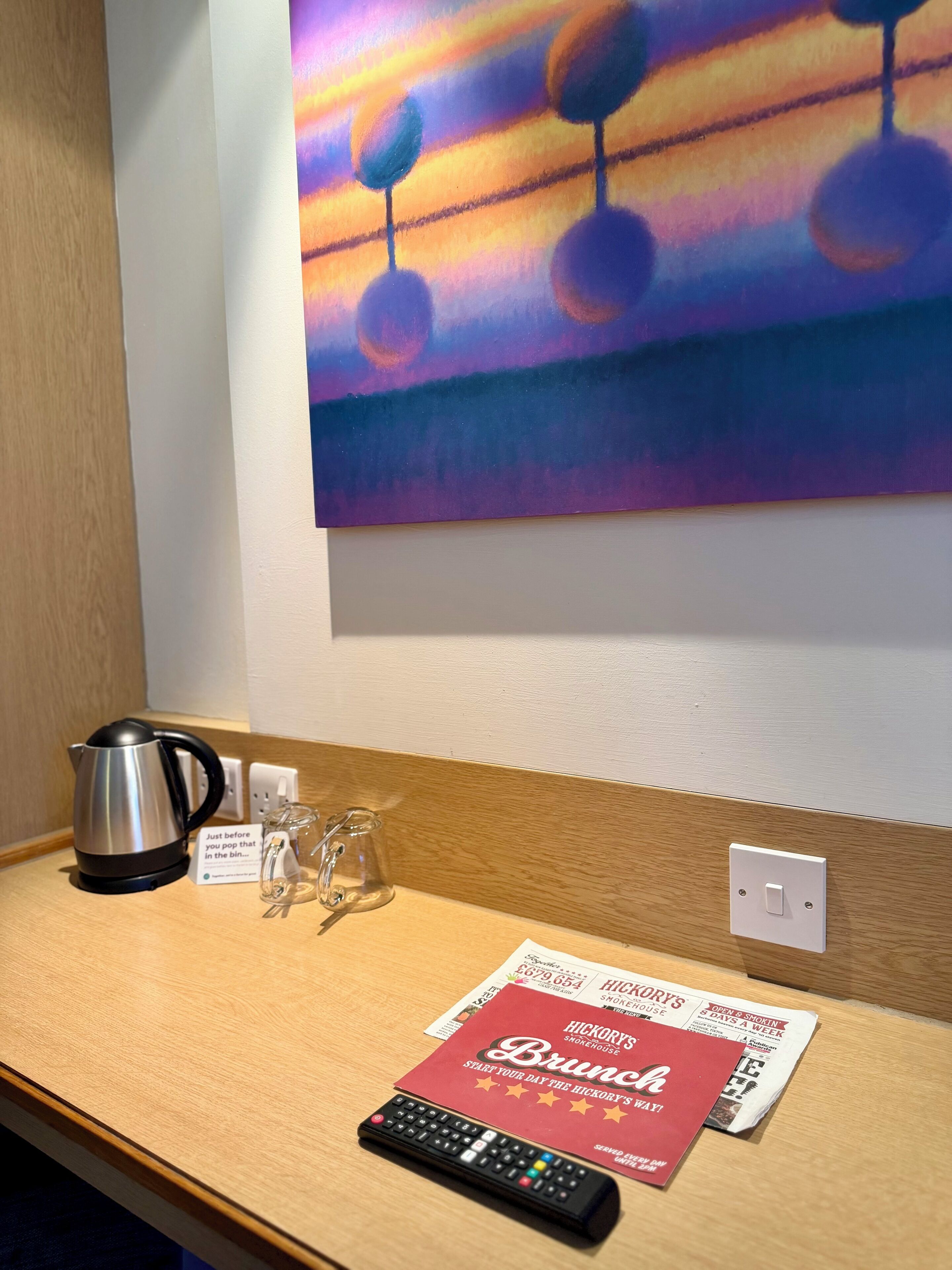 Habitación de hotel con una mesita de madera, una tetera, un vaso, un control remoto y un menú en la pared.