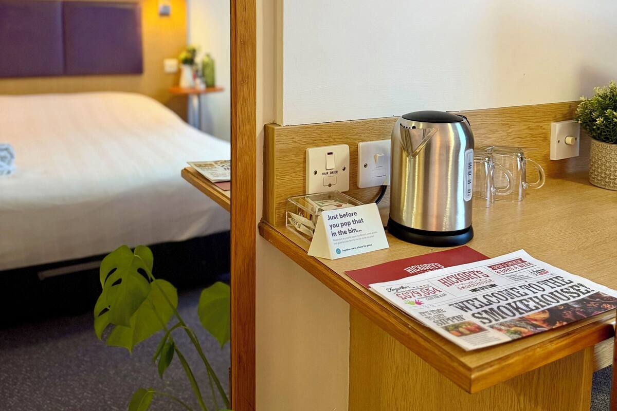 Habitación de hotel con cama, mesita de noche con tetera, un periódico y un cuadro en la pared.