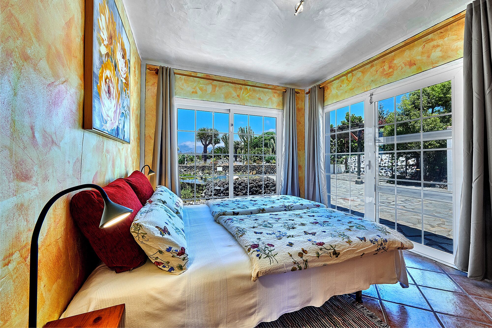 A bedroom with a bed, a nightstand, and a window with a view of a garden.
