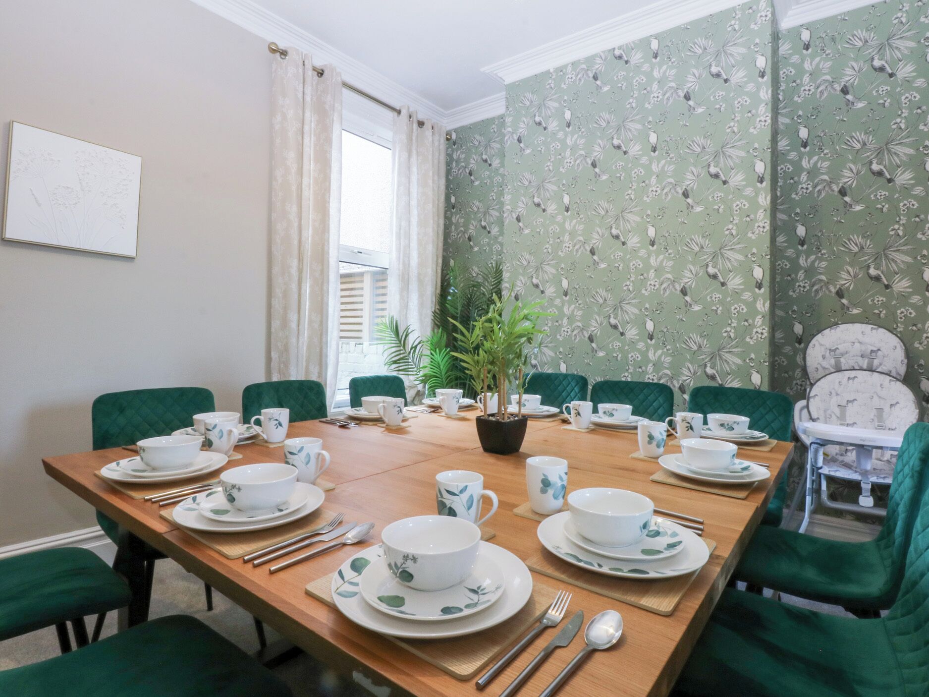 A dining room with a wooden table set for a meal, green upholstered chairs, and a potted plant in the center.