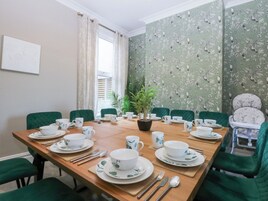A dining room with a wooden table set for a meal, green upholstered chairs, and a potted plant in the center.